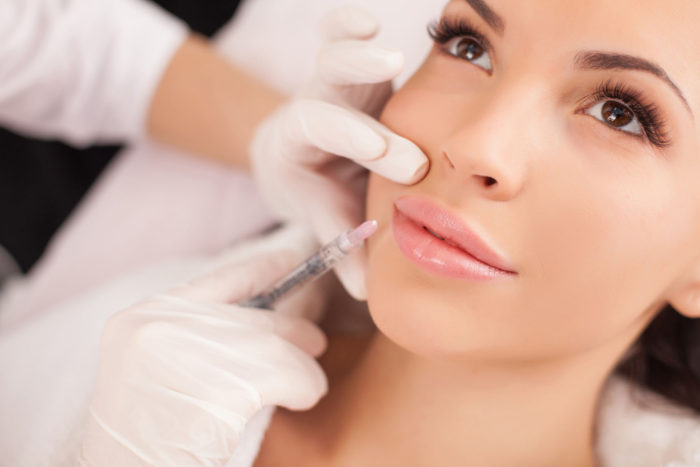 Close up of hands of cosmetologist making botox injection in female lips. She is holding syringe. The young beautiful woman is receiving procedure with enjoyment