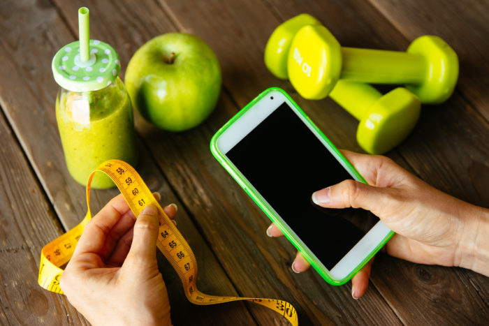 Fitness diet nutrition and workout routine smartphone app concept. Healthy green detox, apple and dumbbells for slimming down dieting. Female checking weight loss with measuring tape.